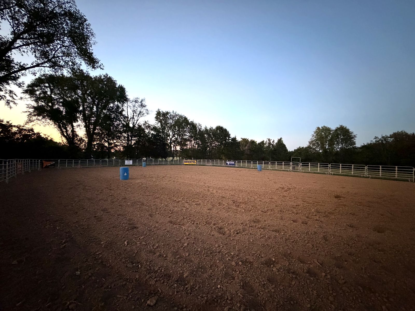 The outdoor dirt arena at dusk, with pipe panel fencing and barrels set out.